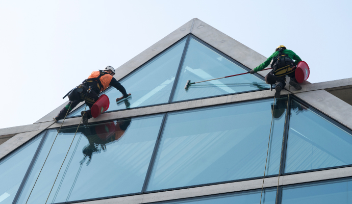 Two workers in safety harnesses cleaning the large glass windows of a commercial building